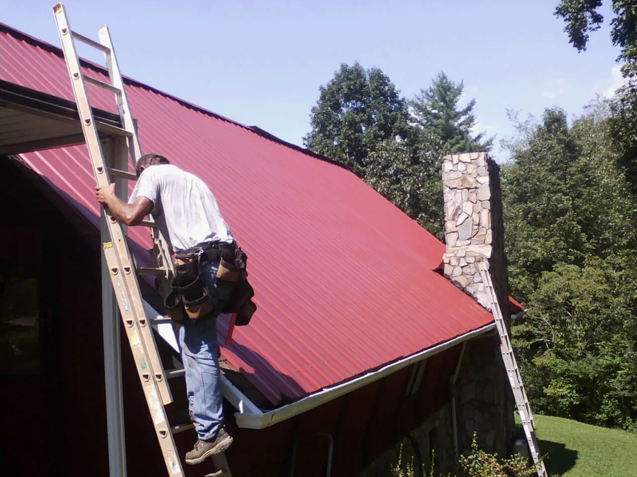 Roofer on a ladder beside a red metal roof and stone chimney — Soffit & Fascia Repair by Roofing Brothers in Salt Lick